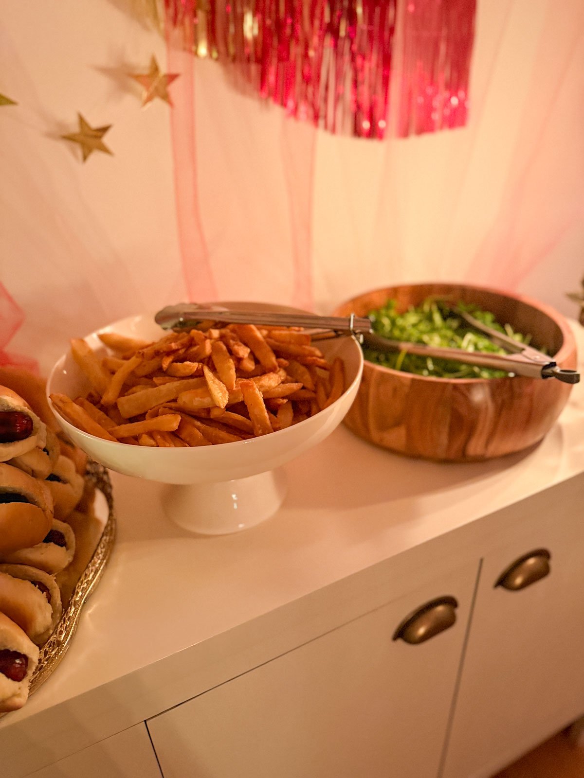 Fresh cut fries in a white pedestal bowl and arugula salad in a wooden bowl on a circus party hot dog buffet, with red and gold tinsel fringe garland and pink tulle backdrop.