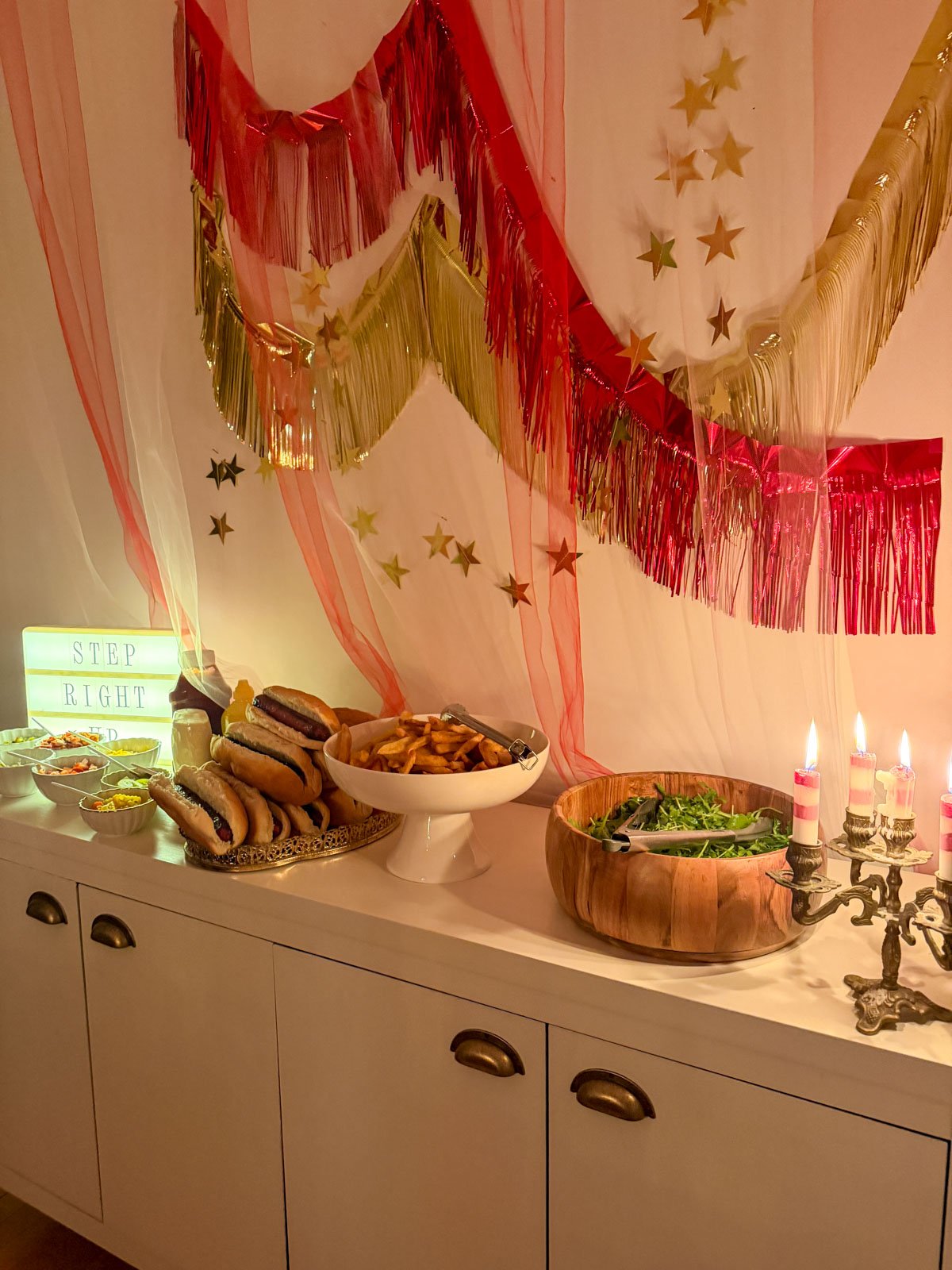 Gourmet hot dog buffet styled against a red and gold tinsel fringe garland backdrop with pink tulle draping, star garlands, a "Step Right Up" lightbox sign, fresh cut fries, and arugula salad at a circus themed dinner party.