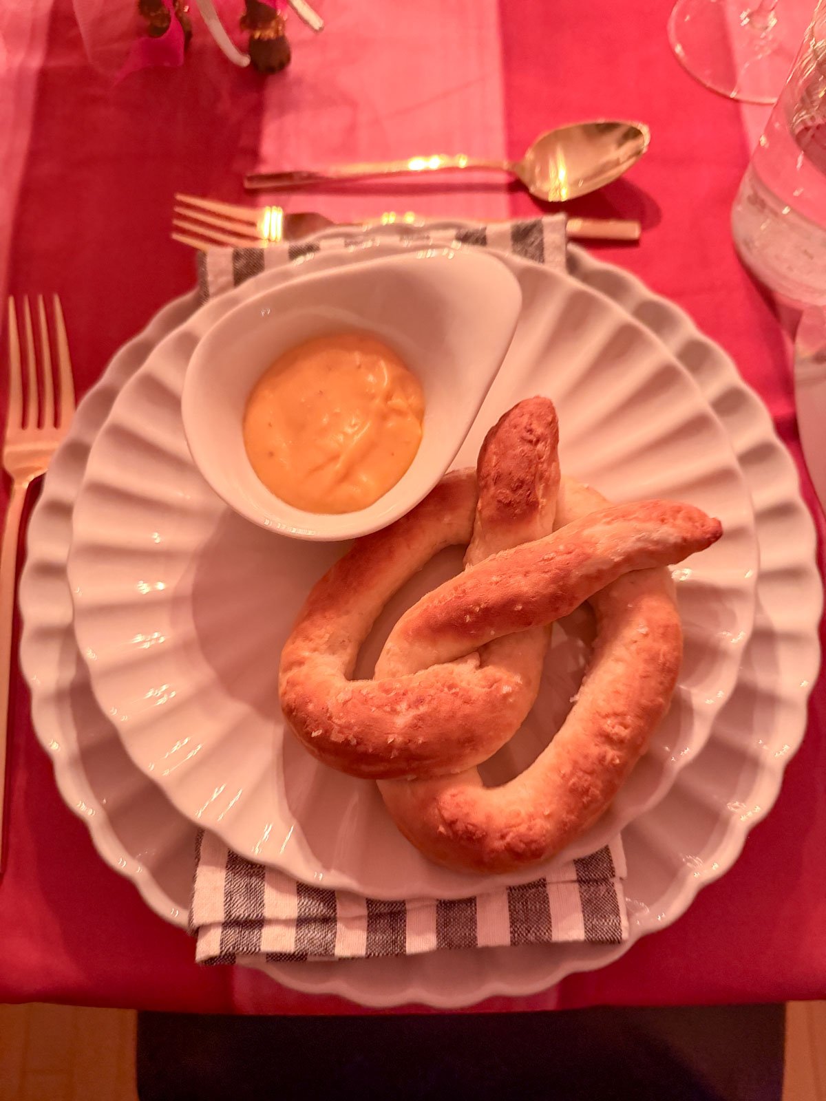 Homemade soft pretzel with beer cheese dipping sauce served on a white scalloped plate at a circus themed dinner party, with gold cutlery and red and pink striped tablecloth.