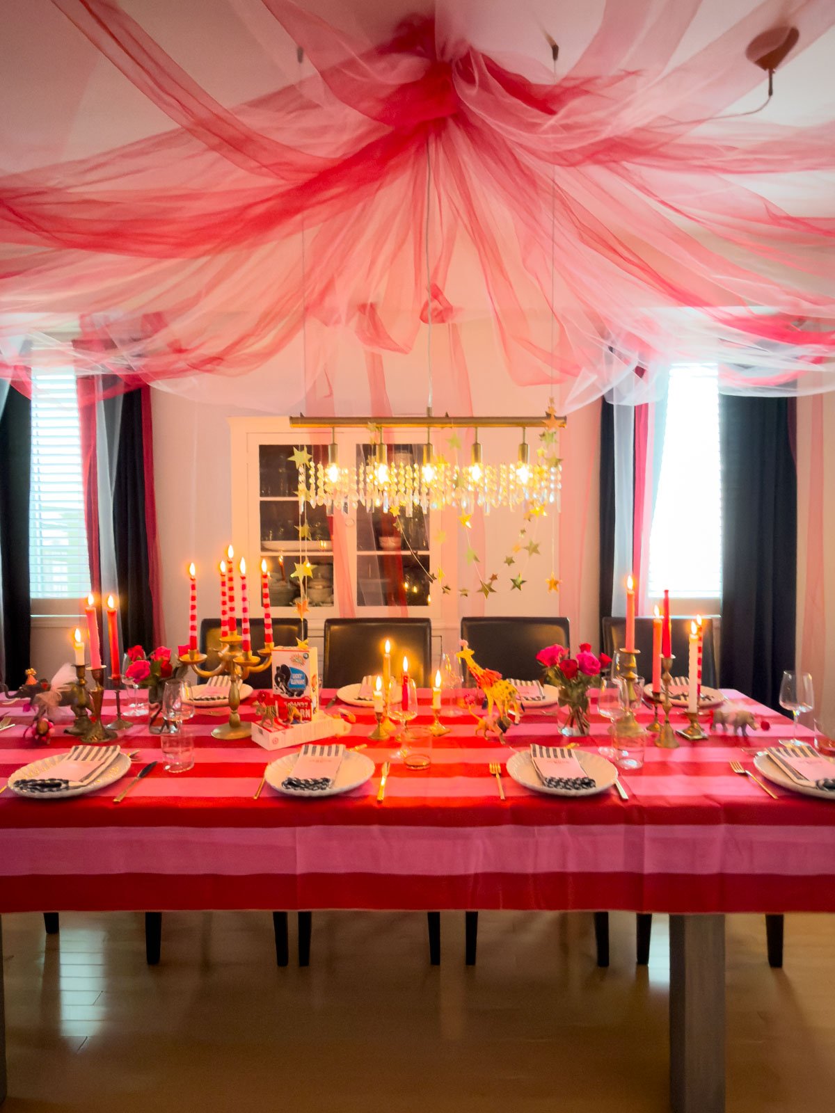 Circus themed dinner party dining room transformed into a big top with a DIY pink and red tulle tent ceiling, fully set table with striped tablecloth, lit brass candlesticks, dressed circus animal figurines, pink roses, and gold star garlands cascading from the chandelier.