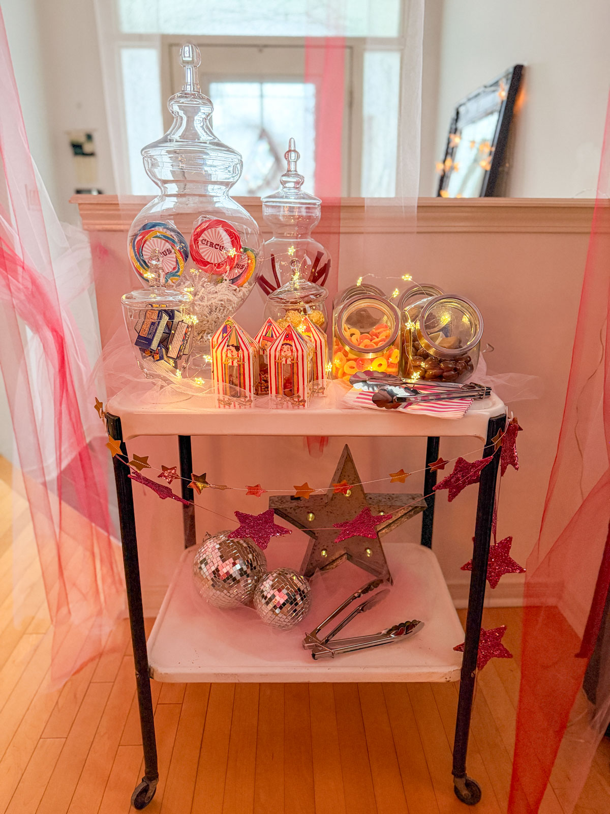 Circus party candy cart styled with glass apothecary jars filled with oversized circus lollipops and sweets, circus tent candy boxes, fairy lights, and pink glitter star garlands, flanked by pink tulle draping.