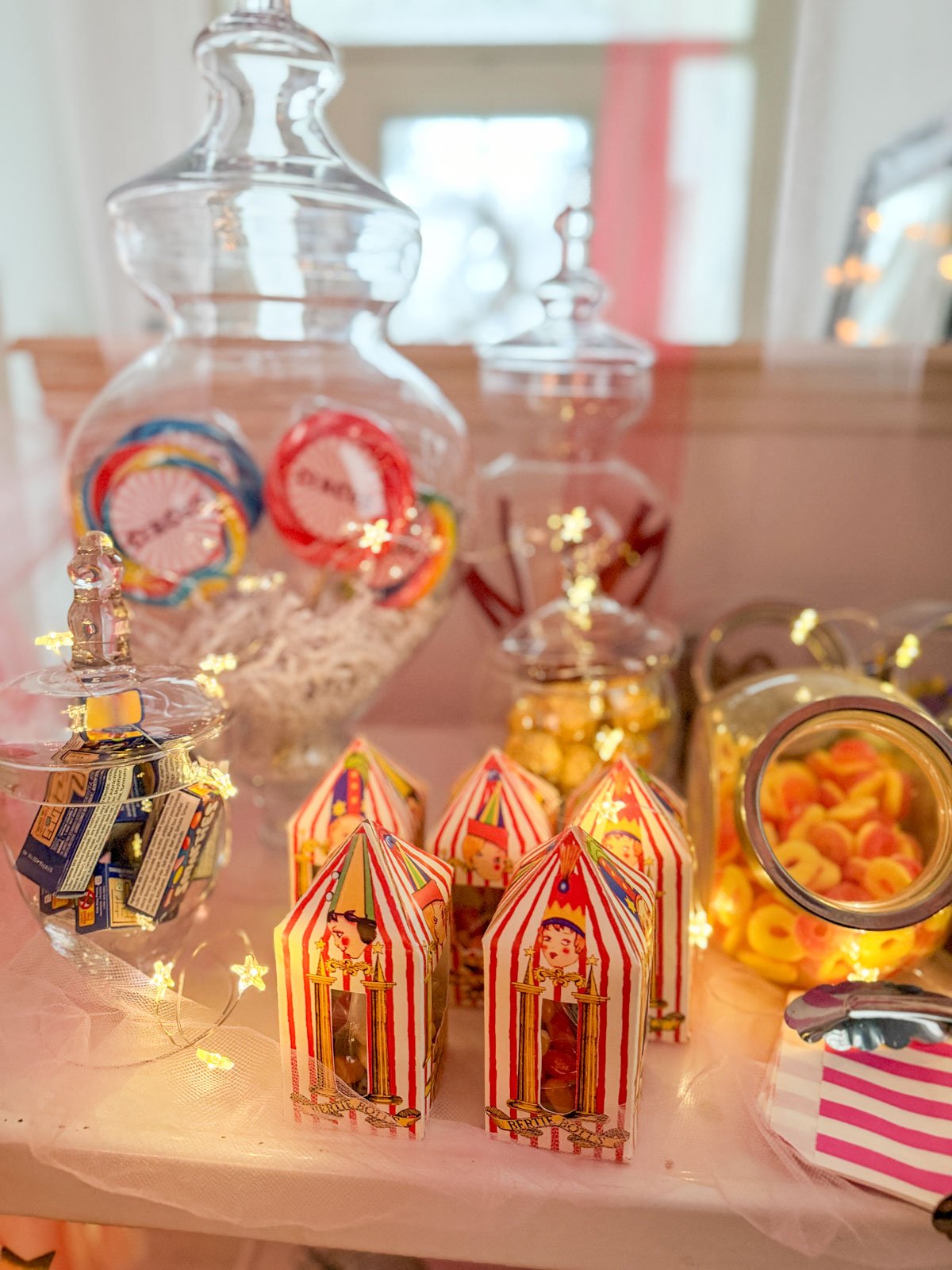 Close-up of circus party candy cart featuring red and white striped circus tent candy boxes, glass apothecary jars filled with oversized circus lollipops and gummy candies, and twinkling star fairy lights on a pink tulle-draped surface.