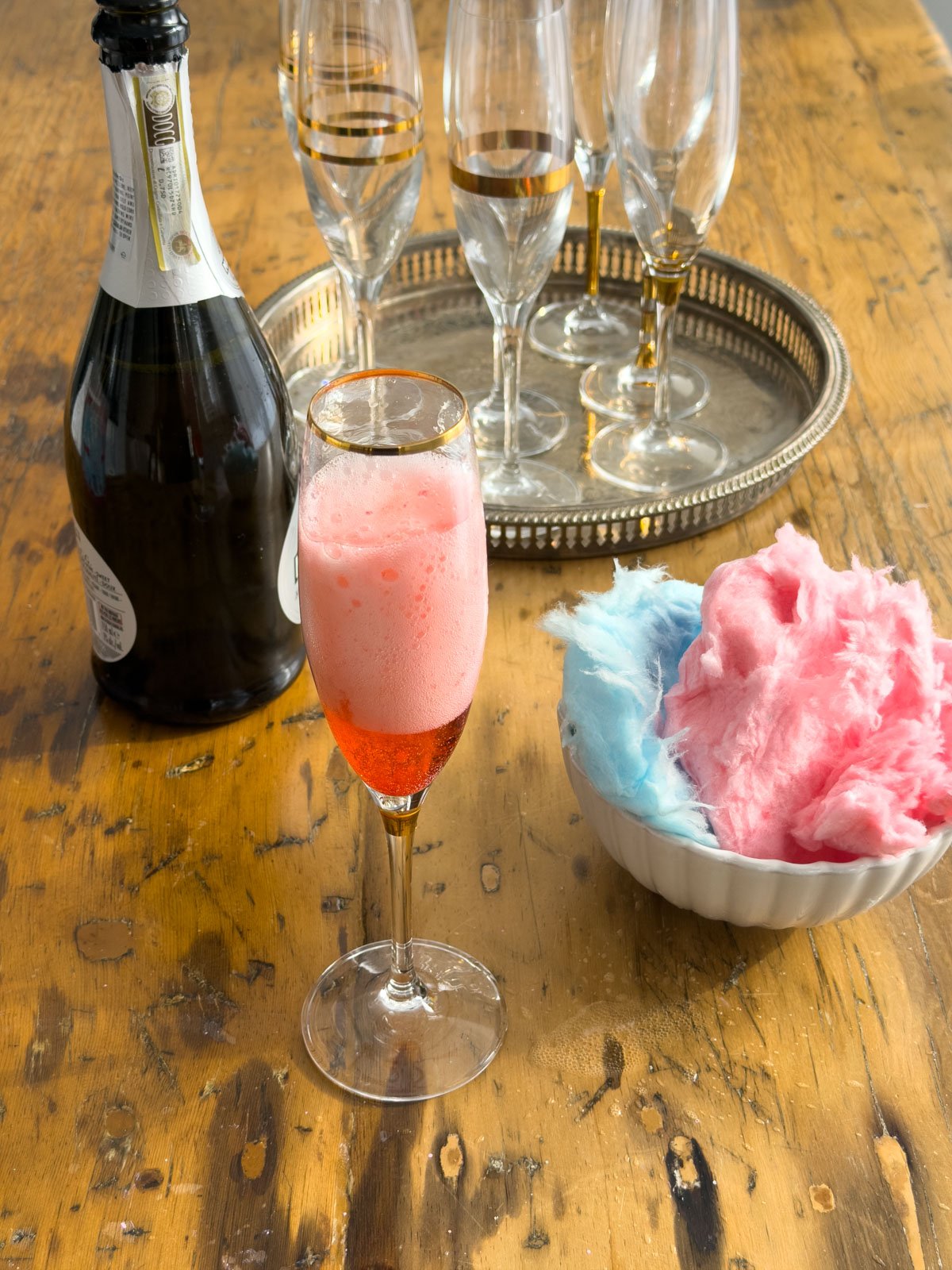 Cotton candy sparkling wine cocktail in a gold-rimmed champagne flute with pink foam, alongside a bowl of pink and blue cotton candy and a bottle of Prosecco on a silver tray with champagne flutes.