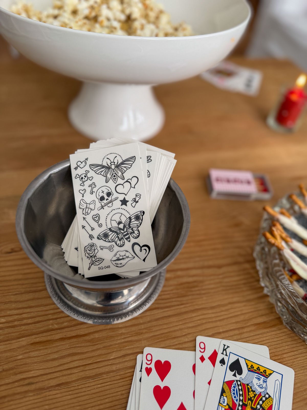 A silver bowl filled with temporary tattoo sheets featuring skull, butterfly, rose, and heart designs, styled on a circus party sideshow coffee table with playing cards, a custom circus matchbox, and chocolate cigarette pretzel rods in the background.