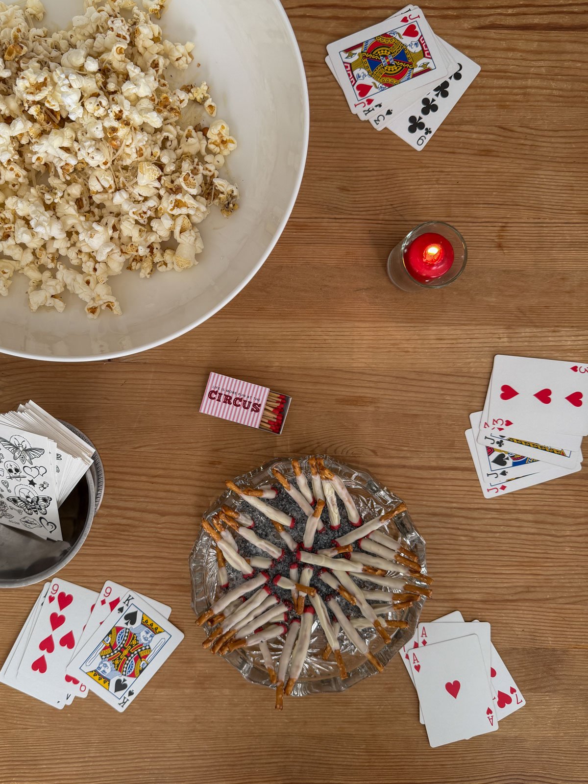 Circus party sideshow coffee table with truffle popcorn, chocolate cigarette pretzel rods in a crystal ashtray, custom circus matchbox, temporary tattoo sheets, and scattered playing cards.