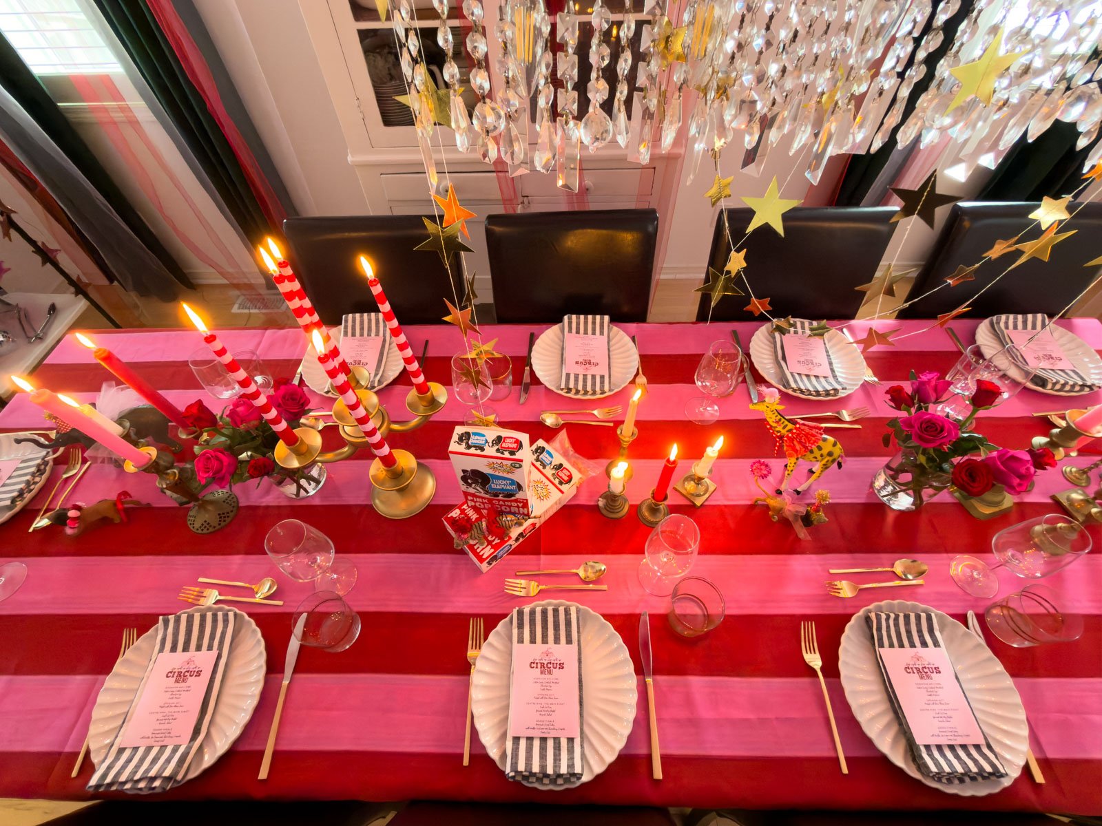 Overhead view of a circus themed dinner party table for eight with a red and pink striped tablecloth, white scalloped plates, striped napkins, gold cutlery, brass candlesticks with lit striped candles, pink roses, dressed circus animal figurines, and gold star garlands.