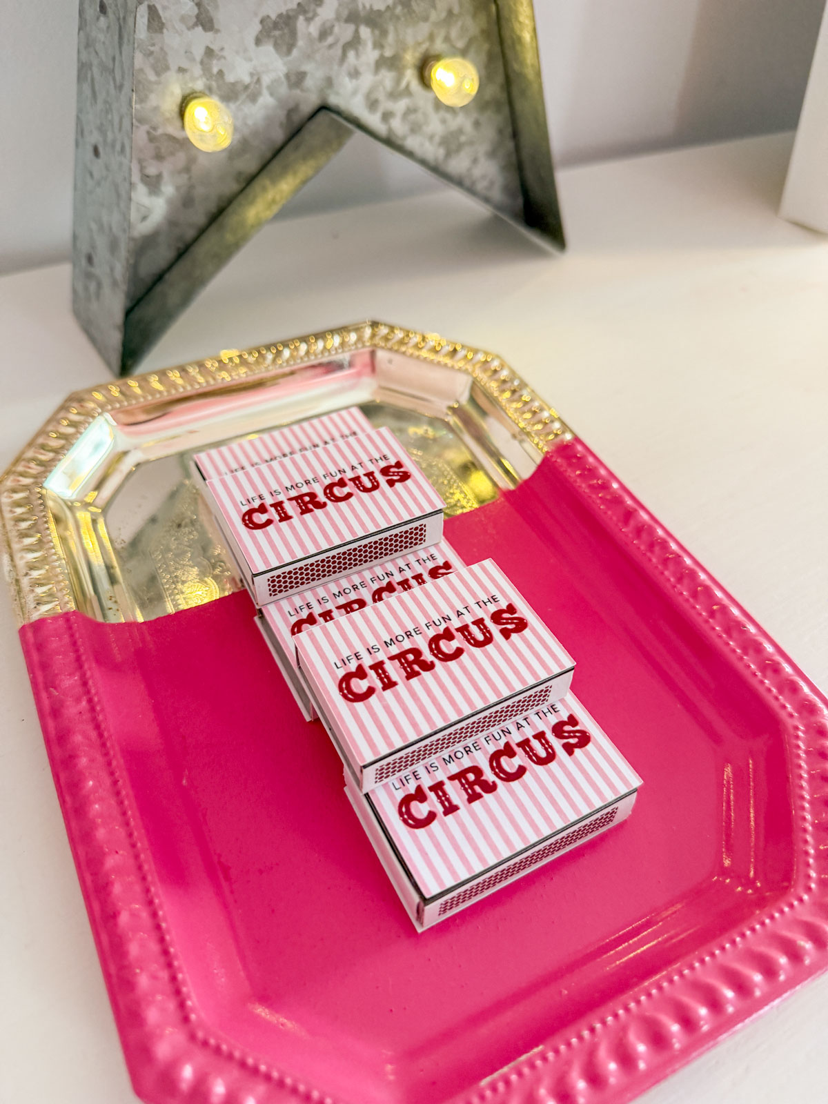 Custom "Life is More Fun at the Circus" DIY matchboxes in pink and white stripes stacked on a hot pink tray with a vintage marquee star light, a circus party favor idea for guests.