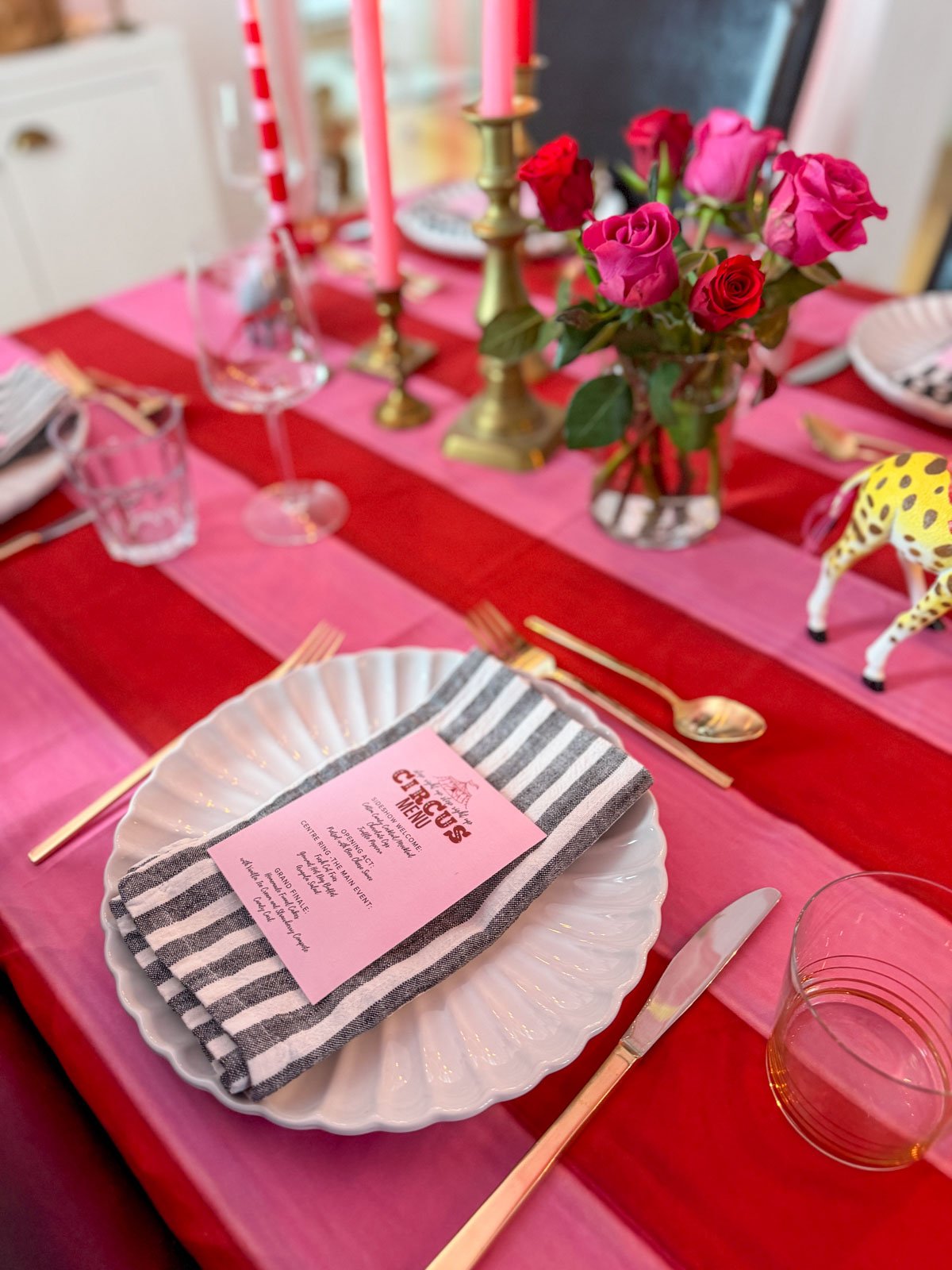 Circus themed dinner party table setting with white scalloped plates, black and white striped napkins, pink Circus Menu cards, and gold cutlery on a red and pink striped tablecloth, with brass candlesticks holding pink and striped candles, and hot pink and red roses.