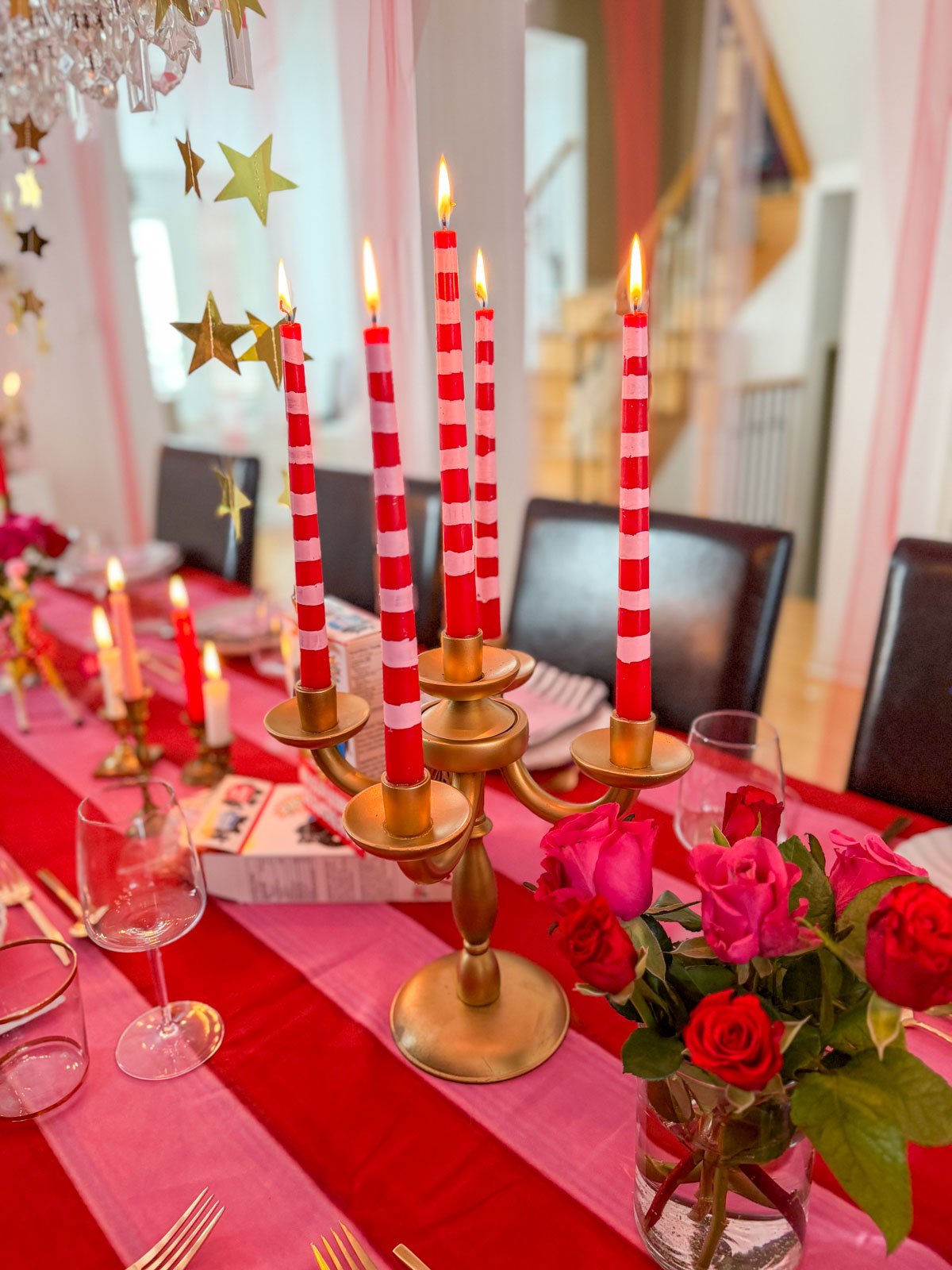 Brass candelabra holding lit hand-painted red and white striped DIY taper candles on a circus themed dinner party table, with hot pink roses, gold star garlands, and the full table setting stretching into the background.