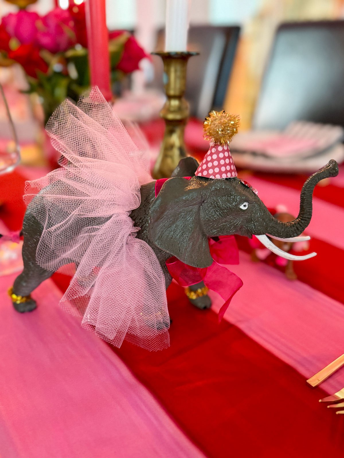 DIY dressed circus elephant figurine wearing a pink tulle skirt, party hat, and gold anklets on a circus themed dinner party table with brass candlesticks and hot pink roses.