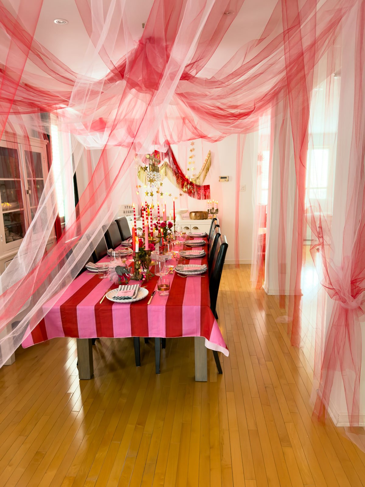 Circus party ideas for adults featuring a DIY tulle tent ceiling over a dressed dinner table with striped candles, brass candlesticks and red and pink roses.