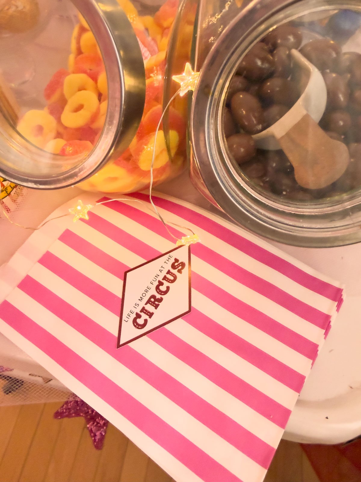 Pink and white striped circus party candy bags with custom "Life is More Fun at the Circus" label, with glass jars of gummy and chocolate candies and star fairy lights in the background.