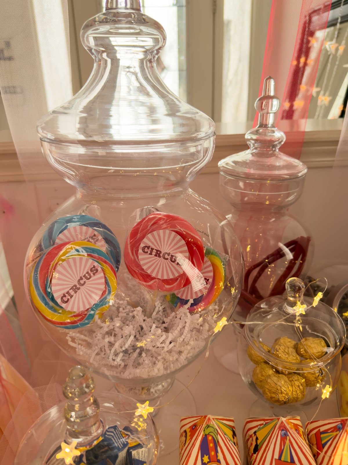 Glass apothecary jar filled with white crinkle paper and oversized circus-labeled lollipops on a circus party candy cart, surrounded by star fairy lights and circus tent candy boxes.