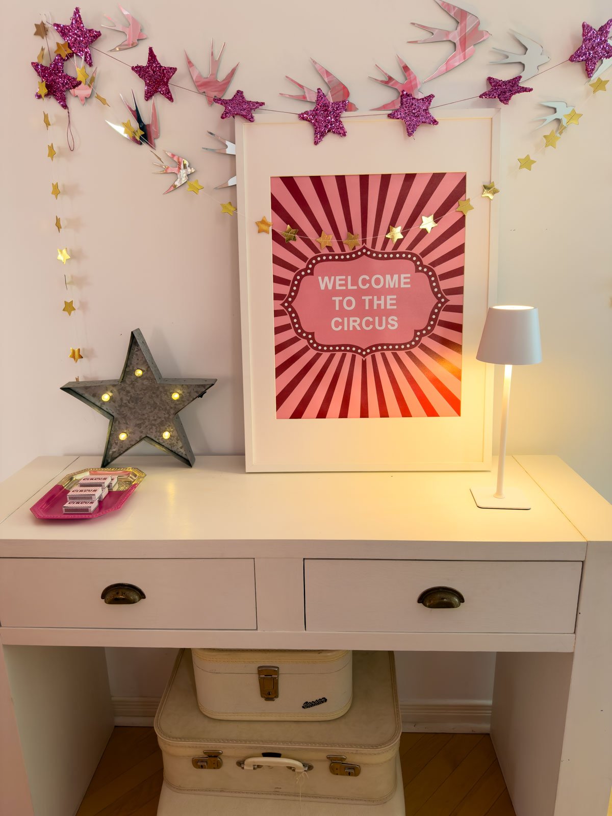 Circus party welcome vignette with a framed "Welcome to the Circus" print, vintage marquee star light, custom matchboxes on a pink tray, and a small lamp on a white console, with swallow and glitter star garlands above and vintage suitcases below.