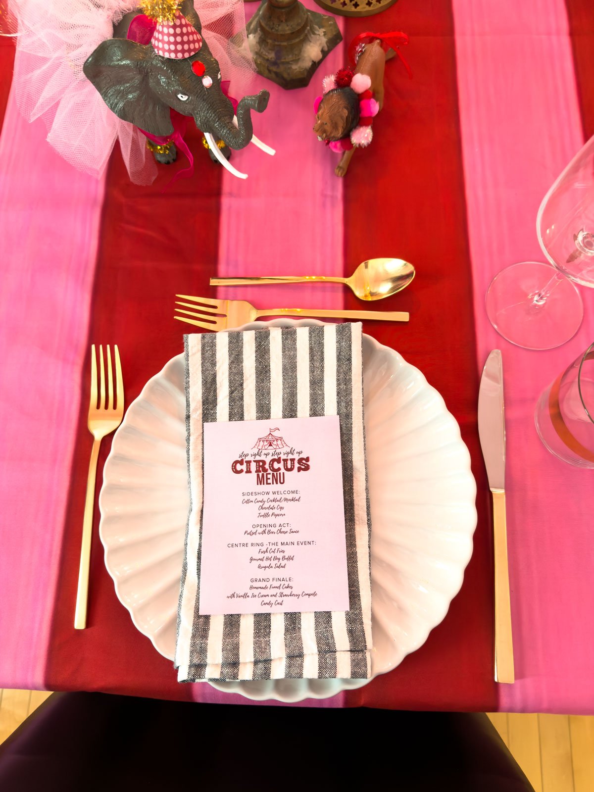 Circus party place setting with a white scalloped plate, black and white striped napkin, pink Circus Menu card, and gold cutlery on a red and pink striped tablecloth, with dressed circus animal figurines in the background.