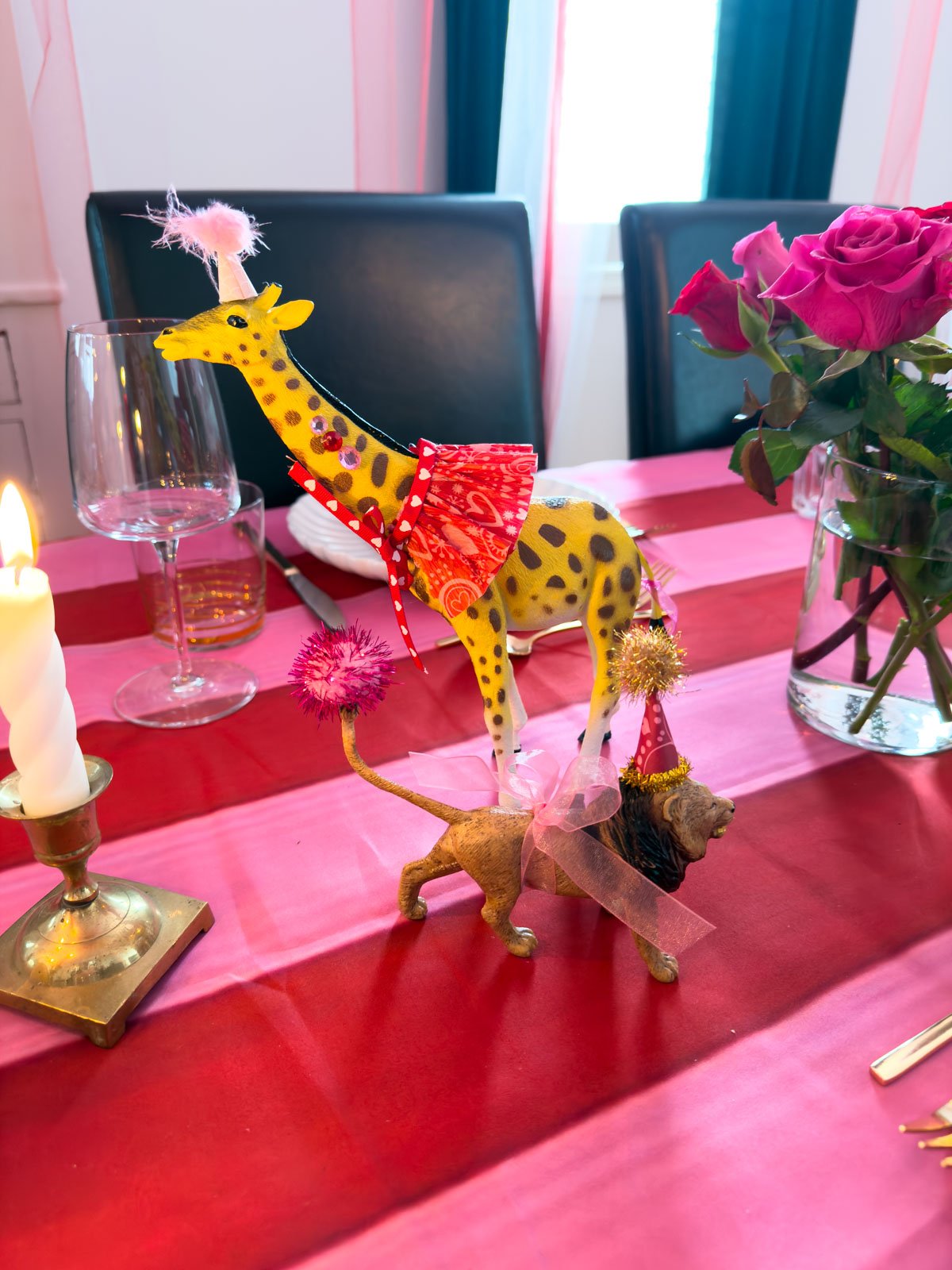 Two dressed circus animal figurines — a giraffe in a ruffled dress and a lion in a gold party hat — on a circus themed dinner party table with a lit candle and pink roses.