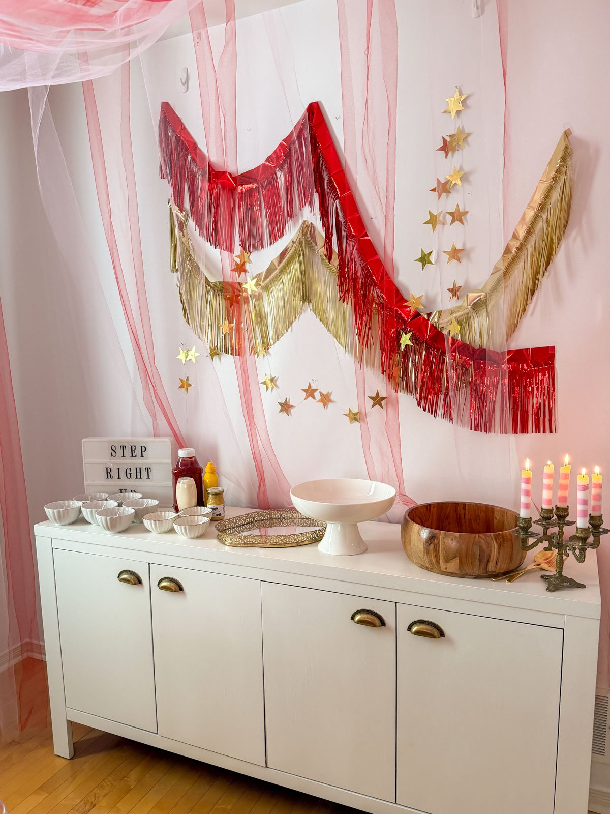 Circus party hot dog buffet backdrop featuring layered red and gold tinsel fringe garlands draped in a V-shape with cascading gold star garlands, pink tulle, a "Step Right Up" lightbox sign, and a brass candelabra with lit striped candles.