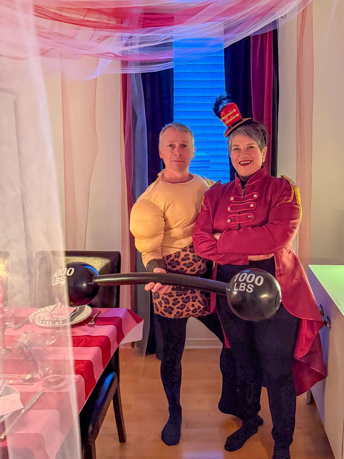 Two circus party guests in costume — one dressed as a strongman in a padded muscle suit holding a 1000 lbs barbell prop, and one as a ringmaster in a red military jacket and feathered top hat — posing under the DIY pink and red tulle big top tent ceiling.