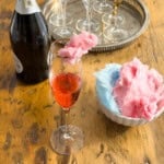 Pink cotton candy cocktail in a gold-rimmed champagne flute with sparkling wine bottle and silver tray of glasses on a wooden table.