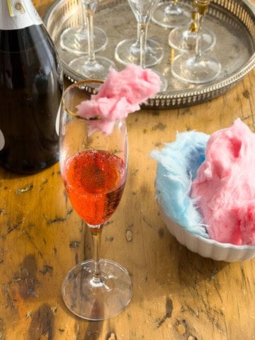 Pink cotton candy cocktail in a gold-rimmed champagne flute with sparkling wine bottle and silver tray of glasses on a wooden table.