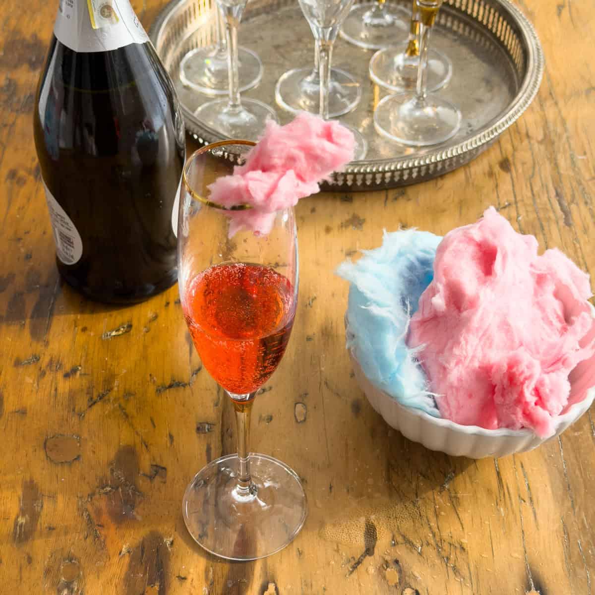 Pink cotton candy cocktail in a gold-rimmed champagne flute with sparkling wine bottle and silver tray of glasses on a wooden table.