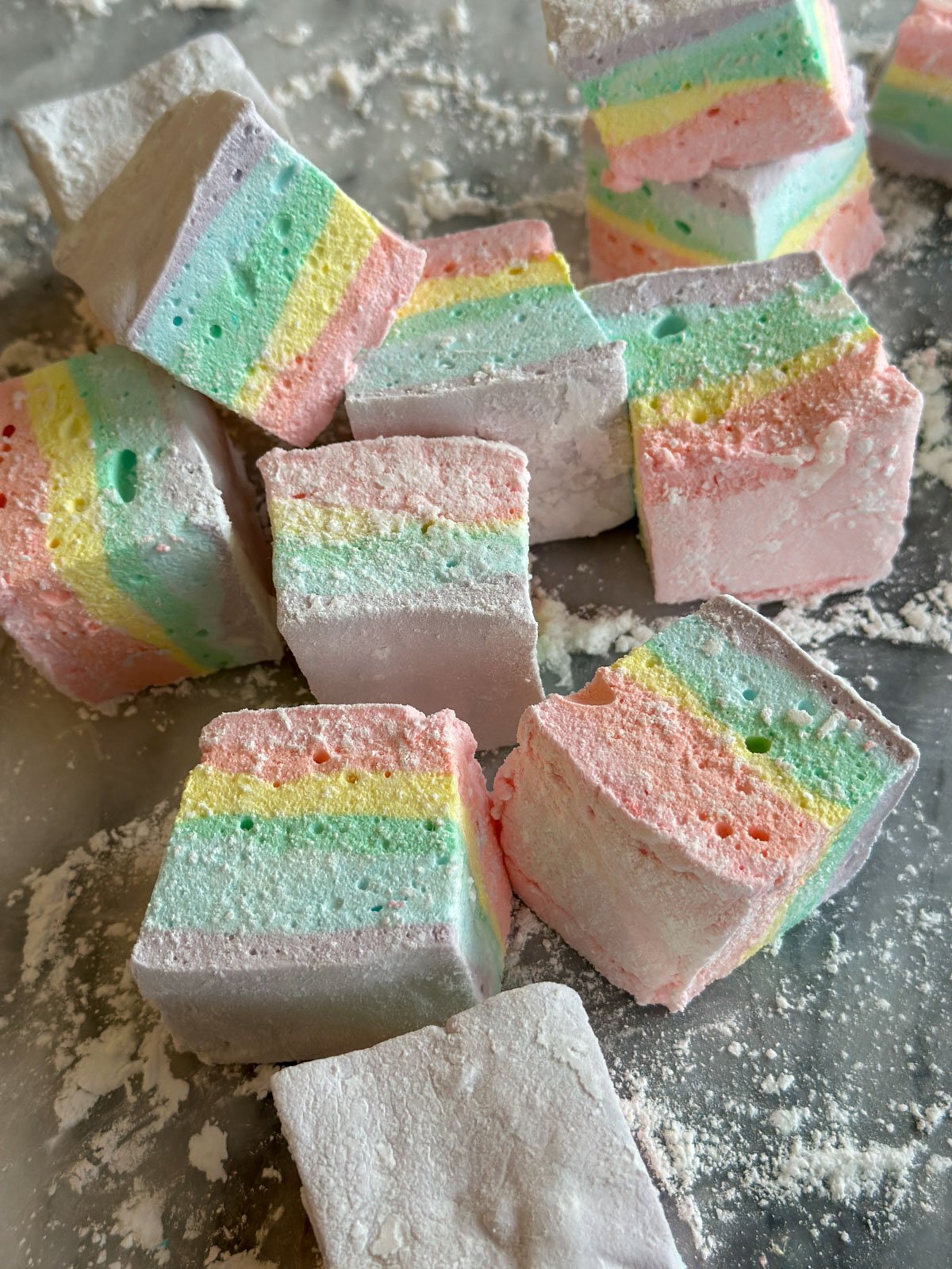 Homemade rainbow marshmallows with pastel layered colors on a marble surface dusted with powdered sugar.