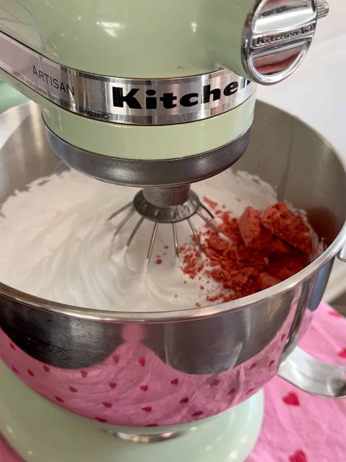 Adding freeze-dried strawberry powder to whipped marshmallow mixture in stand mixer.