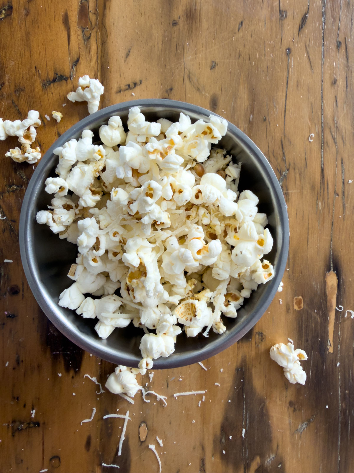 Truffle butter popcorn with freshly grated Parmesan in a silver pewter bowl on a rustic wood surface with scattered popcorn and cheese around the bowl.
