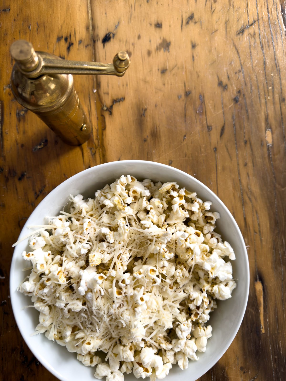 Finished truffle butter popcorn with freshly grated Parmesan and a pinch of cracked pepper in a white bowl, with a vintage brass pepper mill on a rustic wood surface.