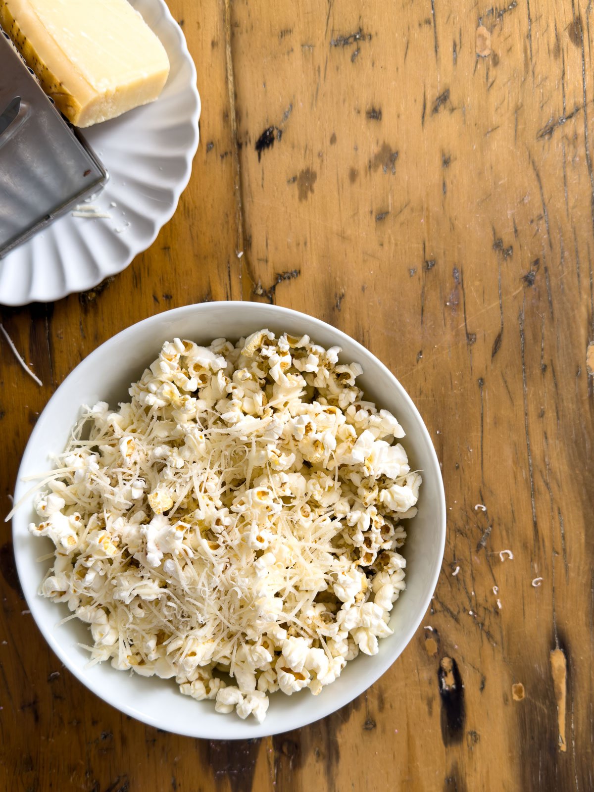 Freshly grated Parmesan being added to truffle butter popcorn in a white bowl, with a box grater and Parmesan wedge on a rustic wood surface.
