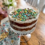 Easter trifle with Cadbury mini eggs on a wooden dining table with a floral napkin and gold spoons.