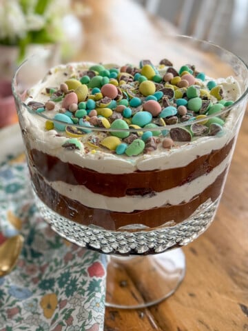 Easter trifle with Cadbury mini eggs on a wooden dining table with a floral napkin and gold spoons.