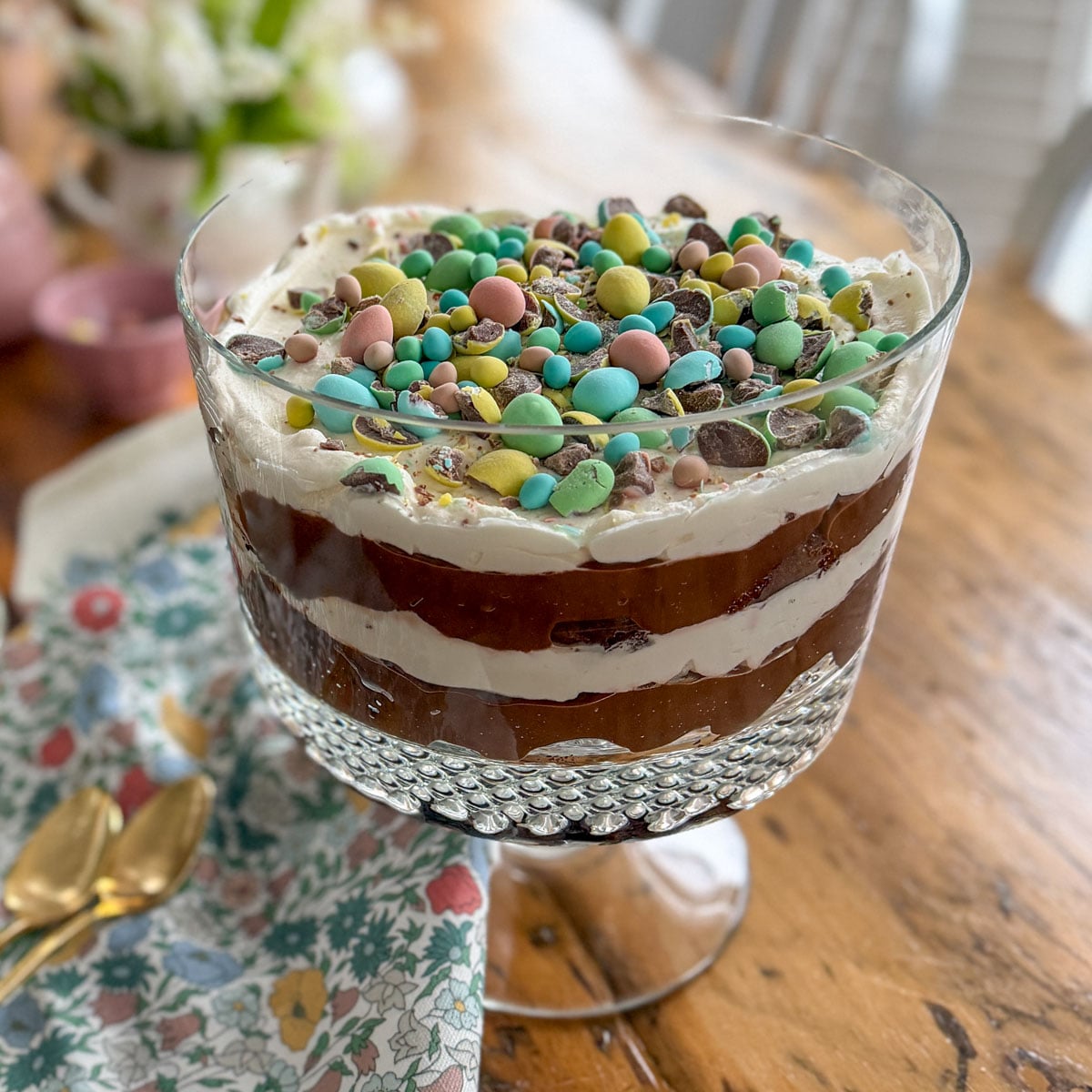 Easter trifle with Cadbury mini eggs on a wooden dining table with a floral napkin and gold spoons.
