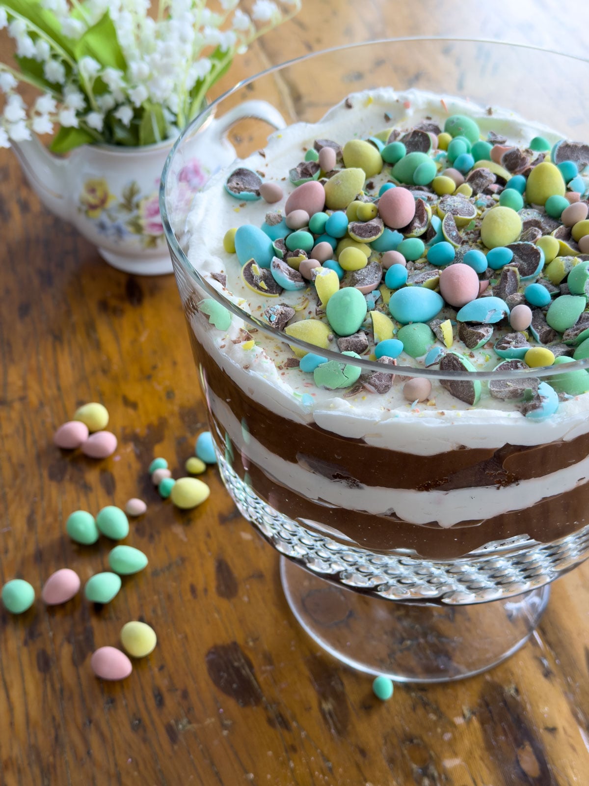Easter trifle with Cadbury mini eggs in a glass trifle bowl with Cadbury mini eggs scattered on a wooden table and lily of the valley flowers in a floral teapot in the background.