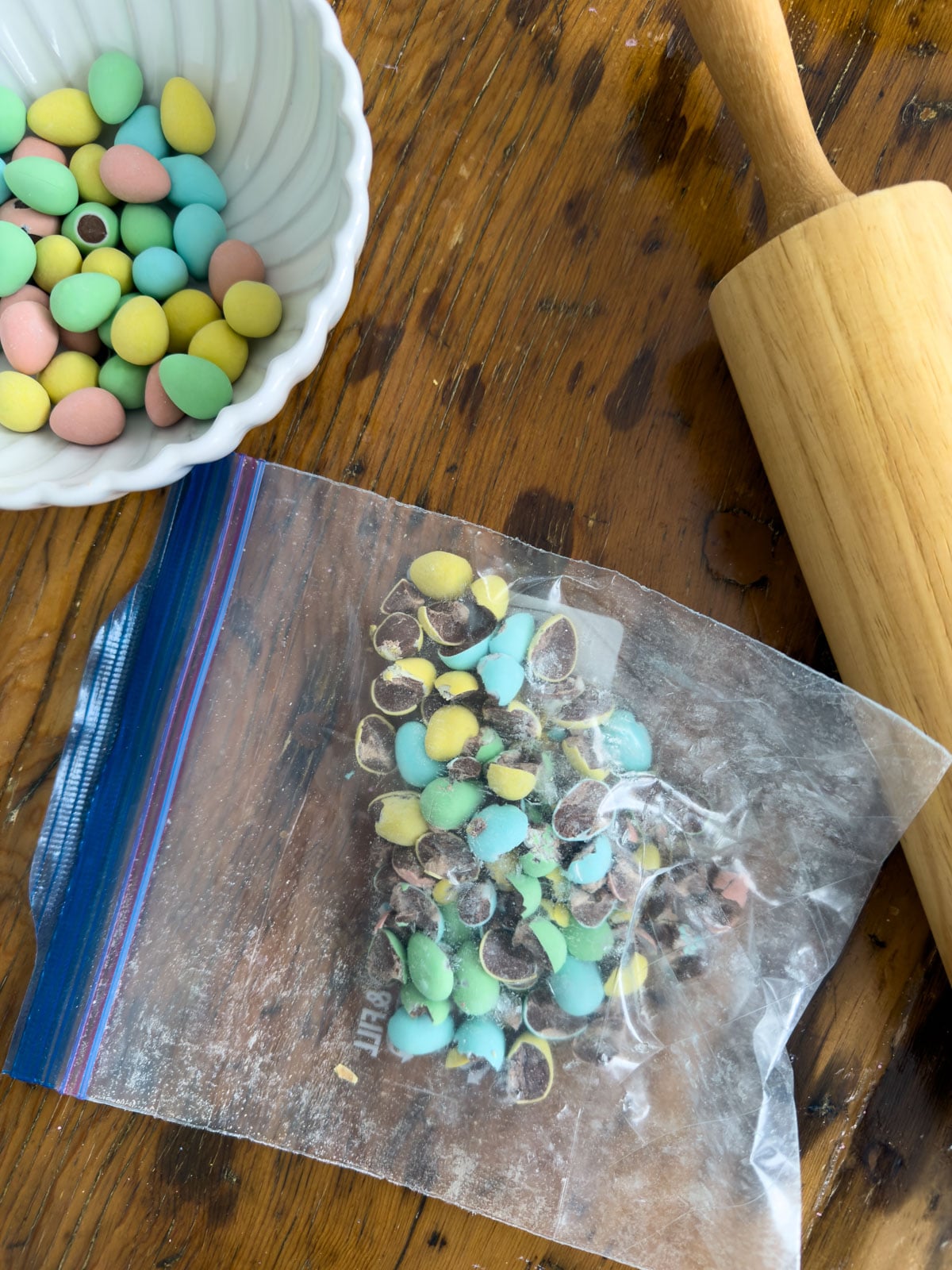 Cadbury mini eggs being broken into pieces inside a zip-lock bag with a wooden rolling pin alongside a bowl of whole mini eggs.