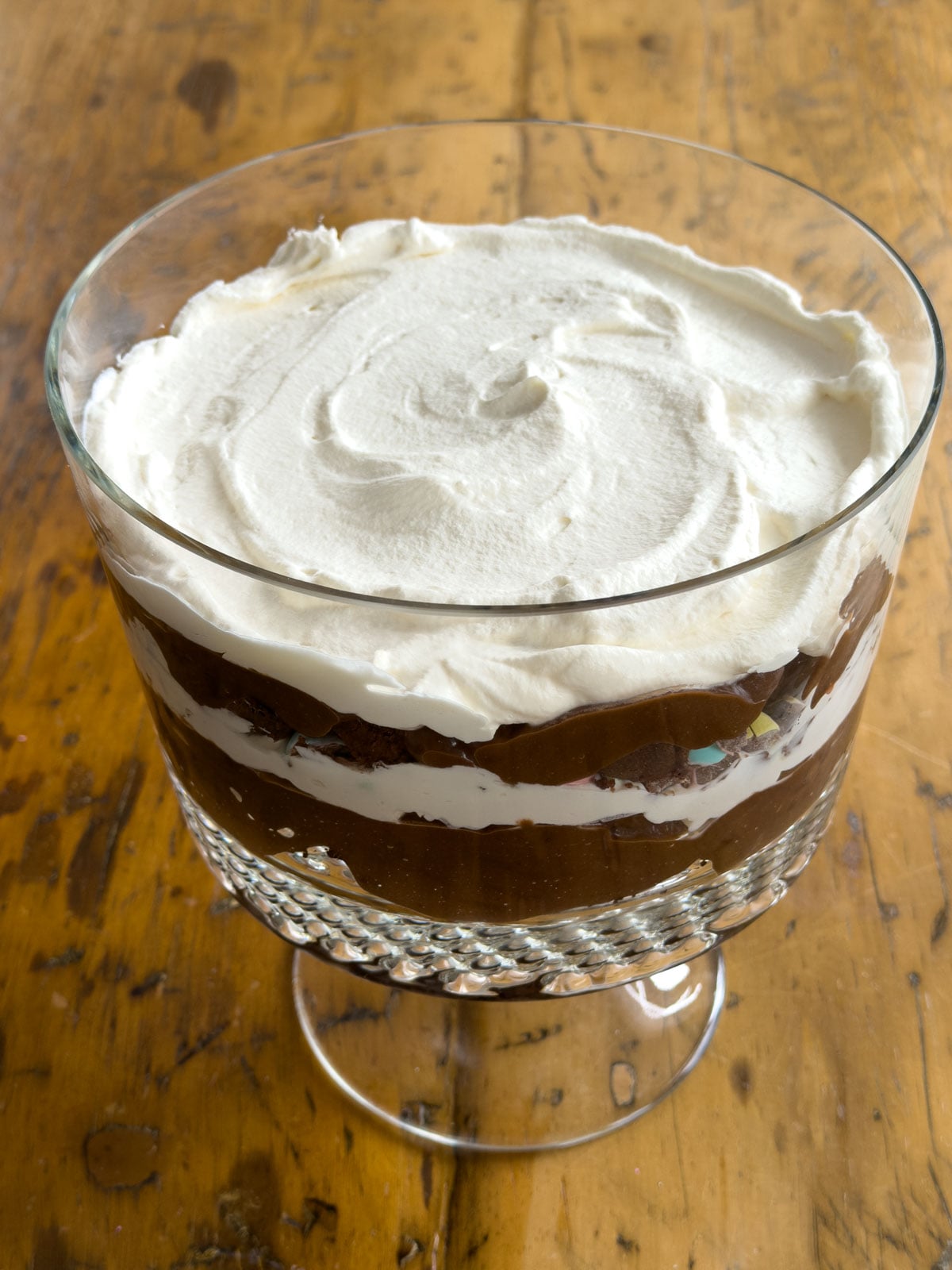 Easter trifle with chocolate pudding and whipped cream layers visible through the glass bowl, ready to chill before adding the mini egg topping.