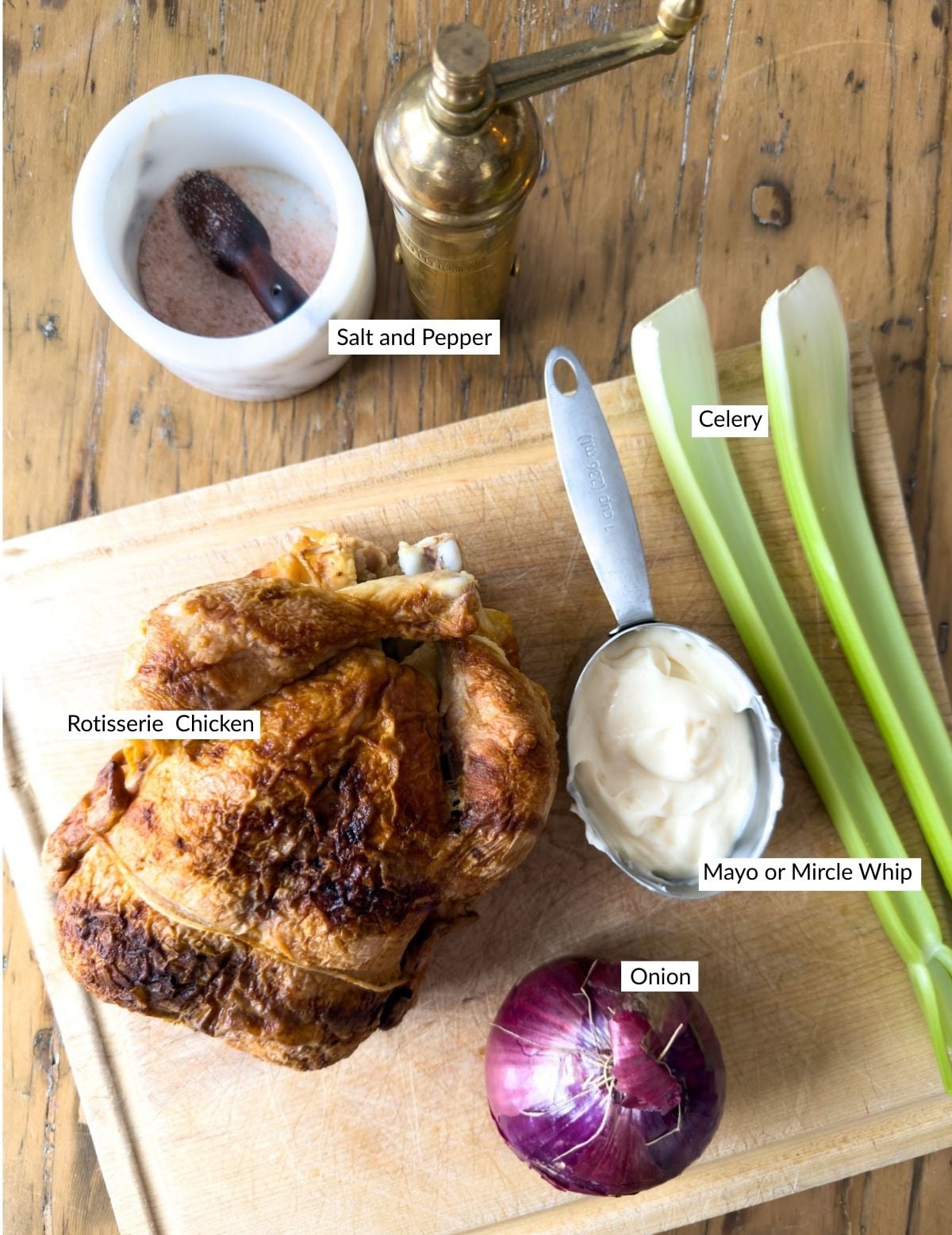 Ingredients for chicken salad finger sandwiches including a whole rotisserie chicken, celery, red onion, mayonnaise, salt and a brass pepper mill on a wooden cutting board.