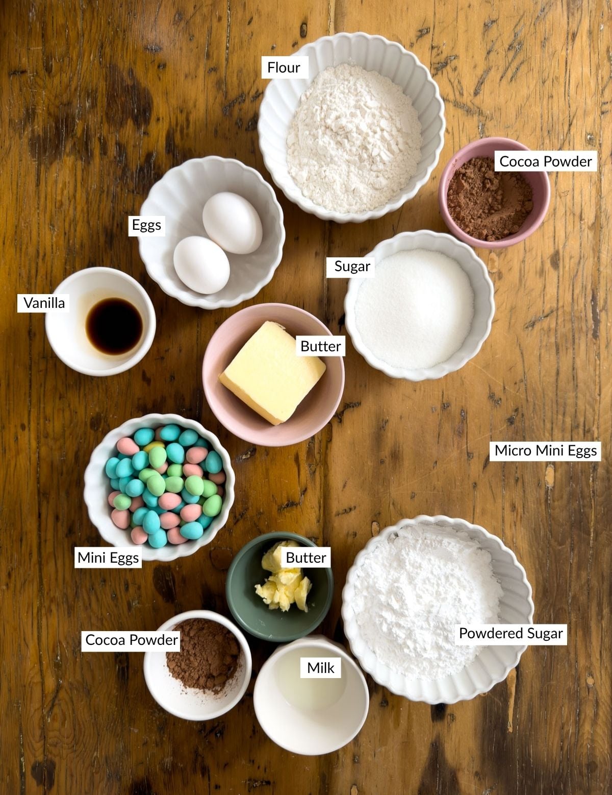 Ingredients for mini egg brownies including flour, eggs, butter, cocoa, sugar and Cadbury mini eggs arranged in small bowls on a wood table.