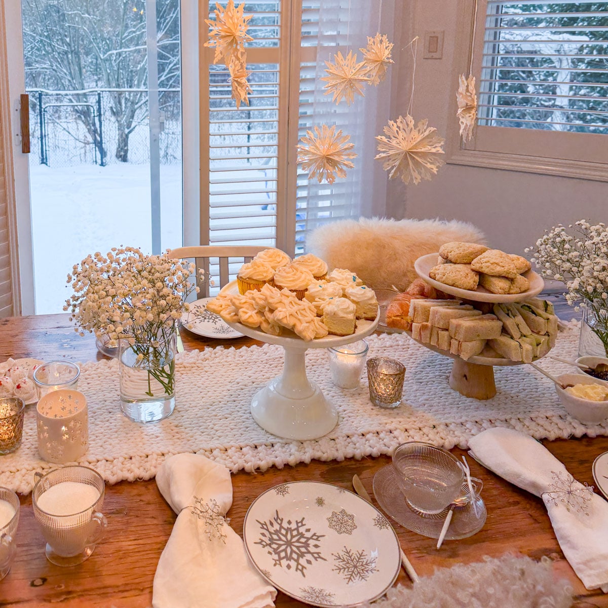 A winter tea party table set with snowflake china, chunky knit runner, and paper snowflake decorations hanging above cake stands filled with frosted cupcakes, scones, and tea sandwiches, with a snowy backyard visible through the windows.