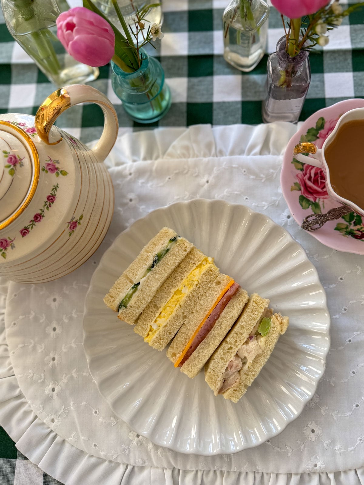 Chicken salad finger sandwiches and assorted tea party finger sandwiches arranged on a white ruffle plate with a rose teapot and pink tulips.