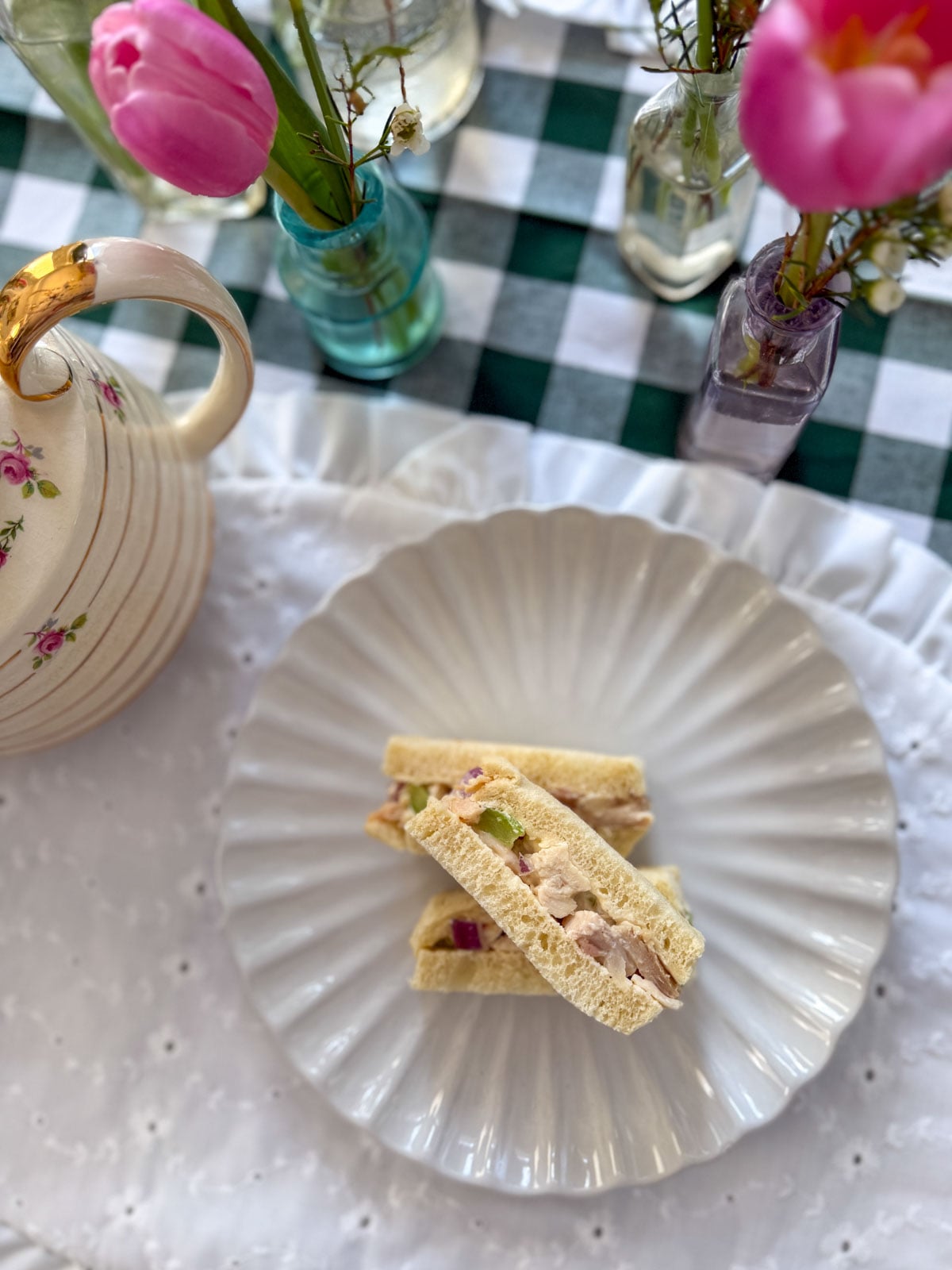 Overhead view of chicken salad finger sandwiches on a white ruffle plate with pink tulips and bud vases on a green gingham tablecloth.
