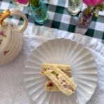 Chicken salad finger sandwiches on a white ruffle plate with a rose teapot and bud vases on a green gingham tablecloth.