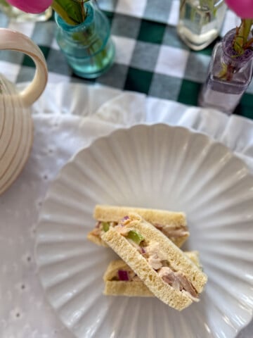 Chicken salad finger sandwiches on a white ruffle plate with a rose teapot and bud vases on a green gingham tablecloth.