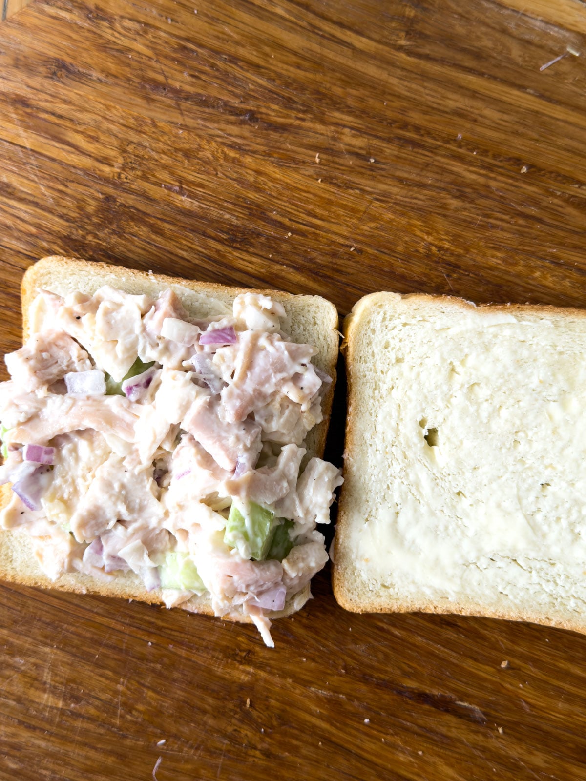Chicken salad spread on one slice of white bread beside a buttered slice during assembly of chicken salad finger sandwiches.