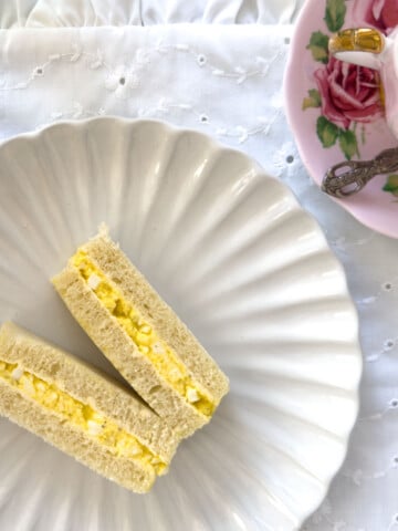 Egg salad finger sandwiches on a white scalloped plate with a rose teacup and saucer on a white eyelet tablecloth.