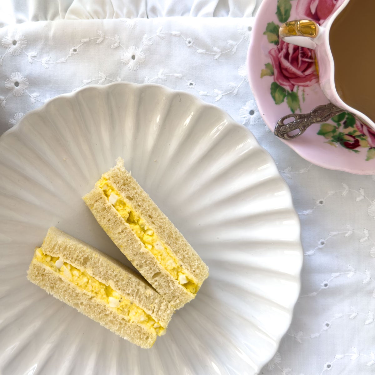 Egg salad finger sandwiches on a white scalloped plate with a rose teacup and saucer on a white eyelet tablecloth.