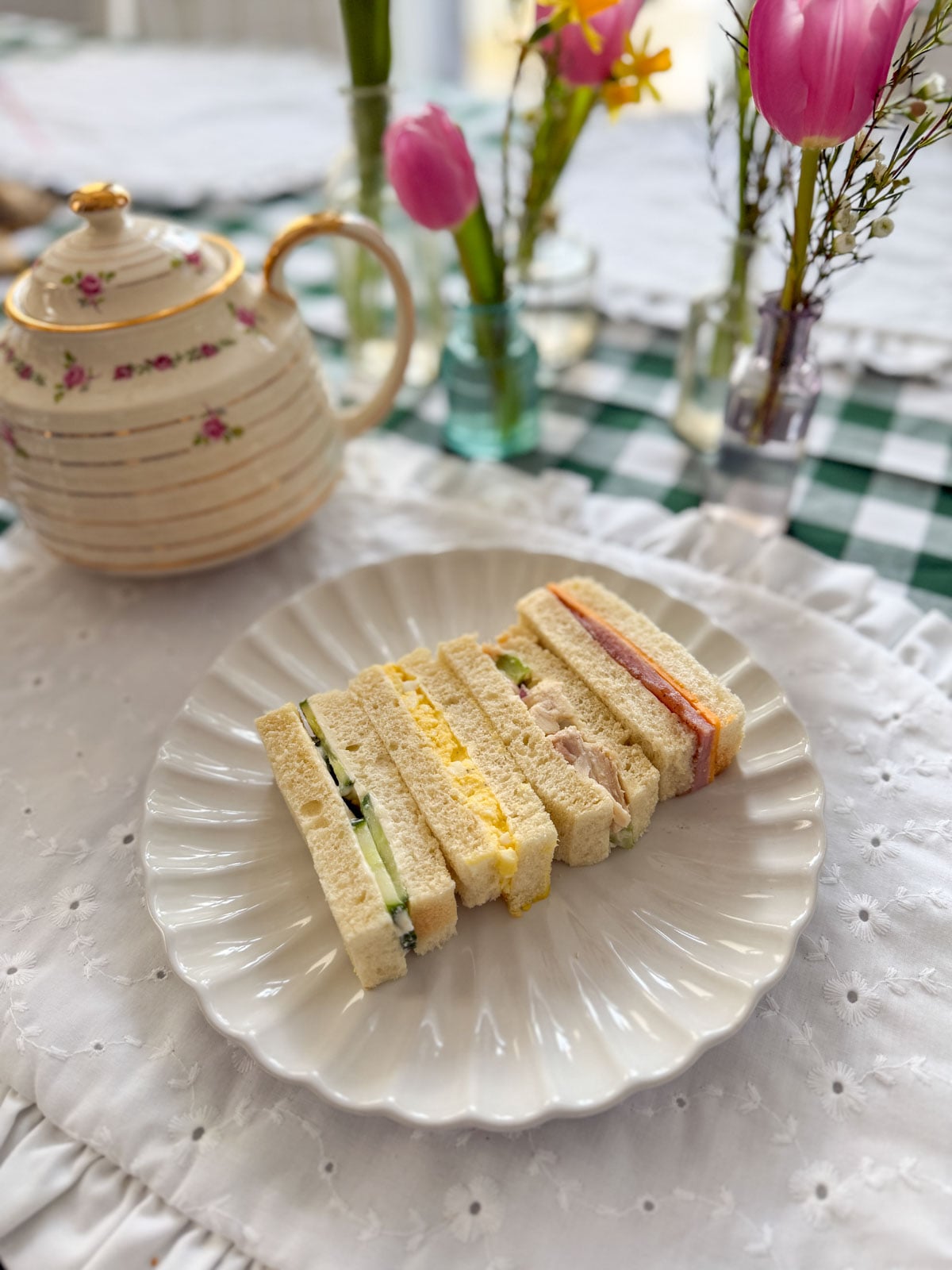 Four varieties of tea party finger sandwiches including egg salad, cucumber, chicken salad, and ham arranged on a white scalloped plate.