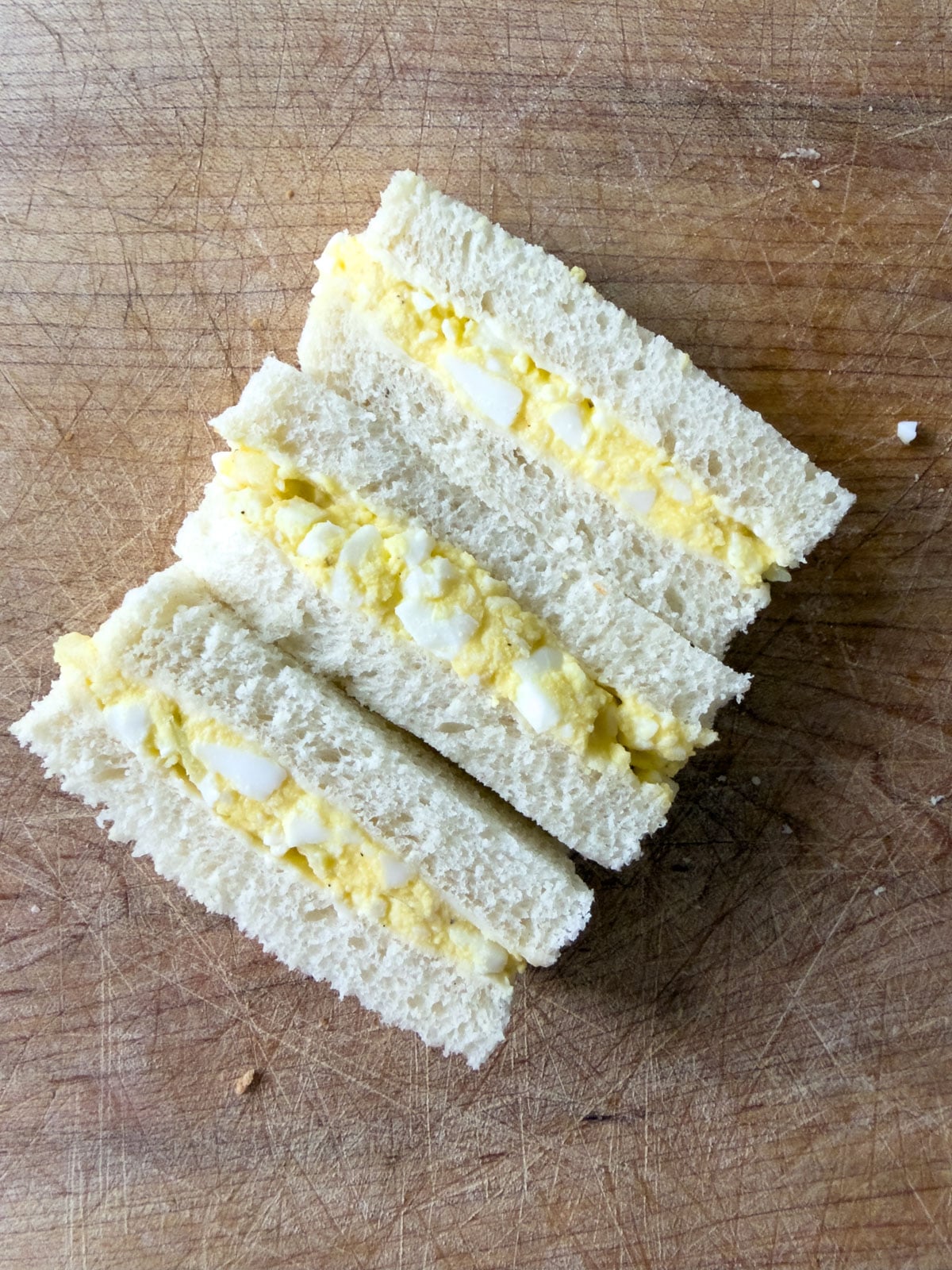 Egg salad finger sandwiches arranged cut side up on a wood cutting board showing the filling.