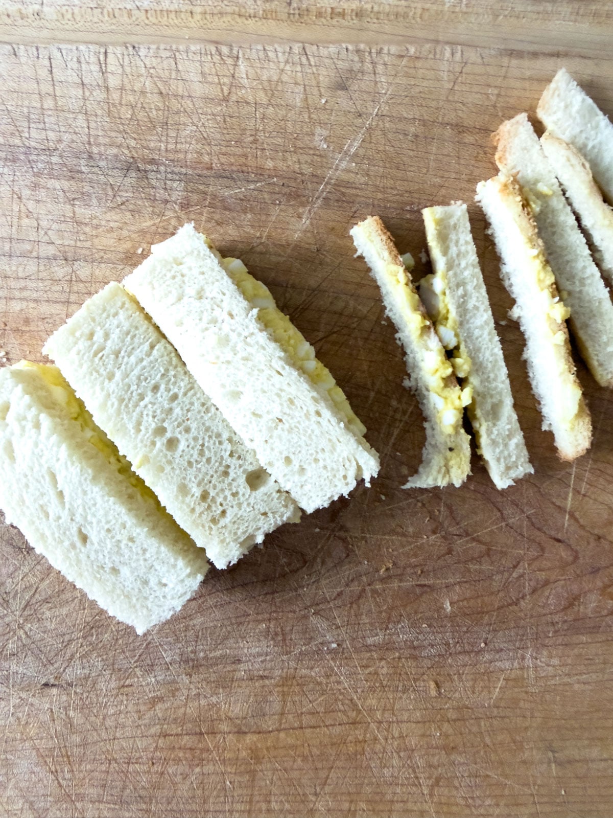 Crustless finger sandwiches on a wood cutting board with the trimmed crusts set to the side.