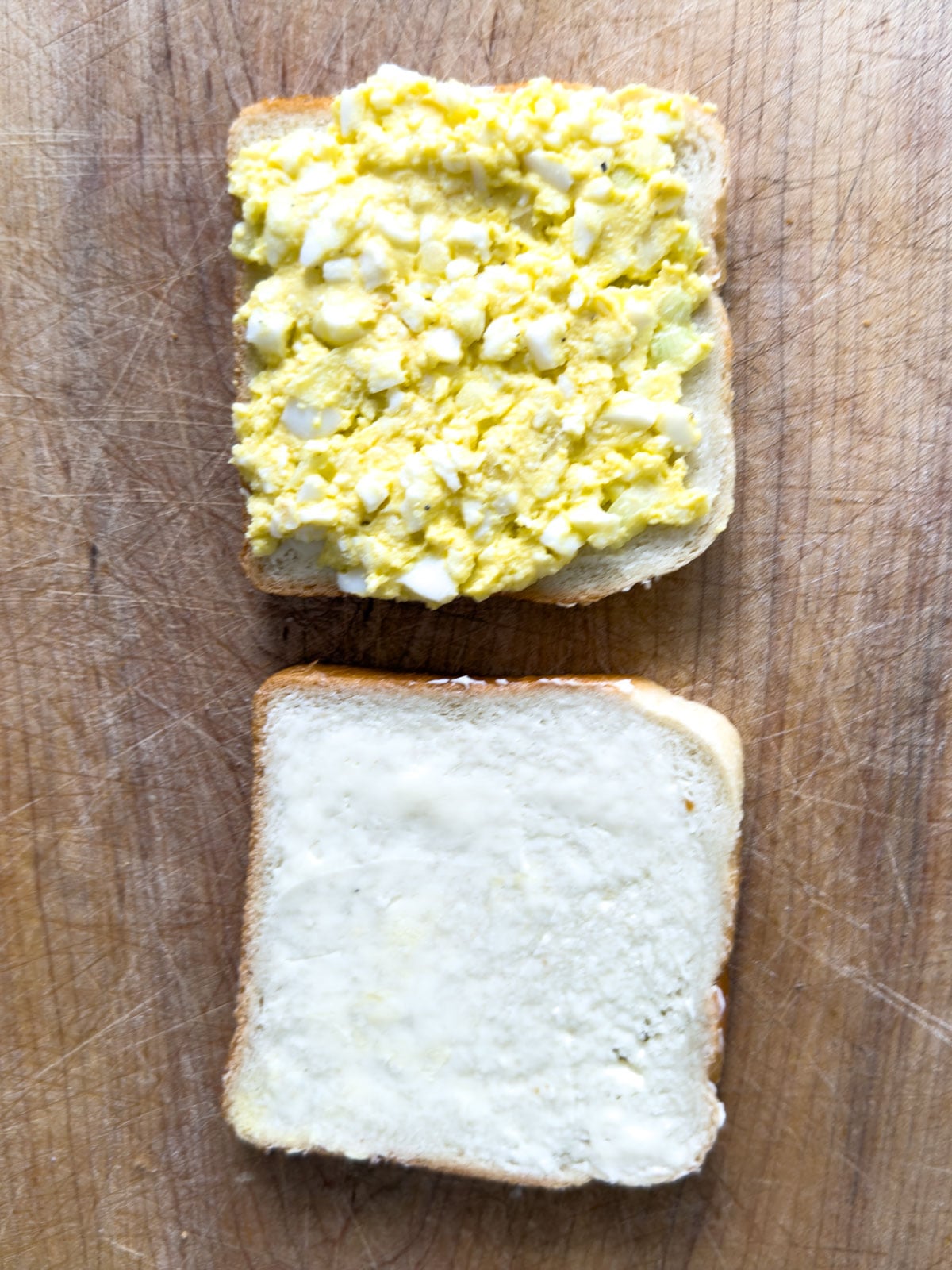 Egg salad spread on a slice of buttered white bread with a second slice ready to top it.